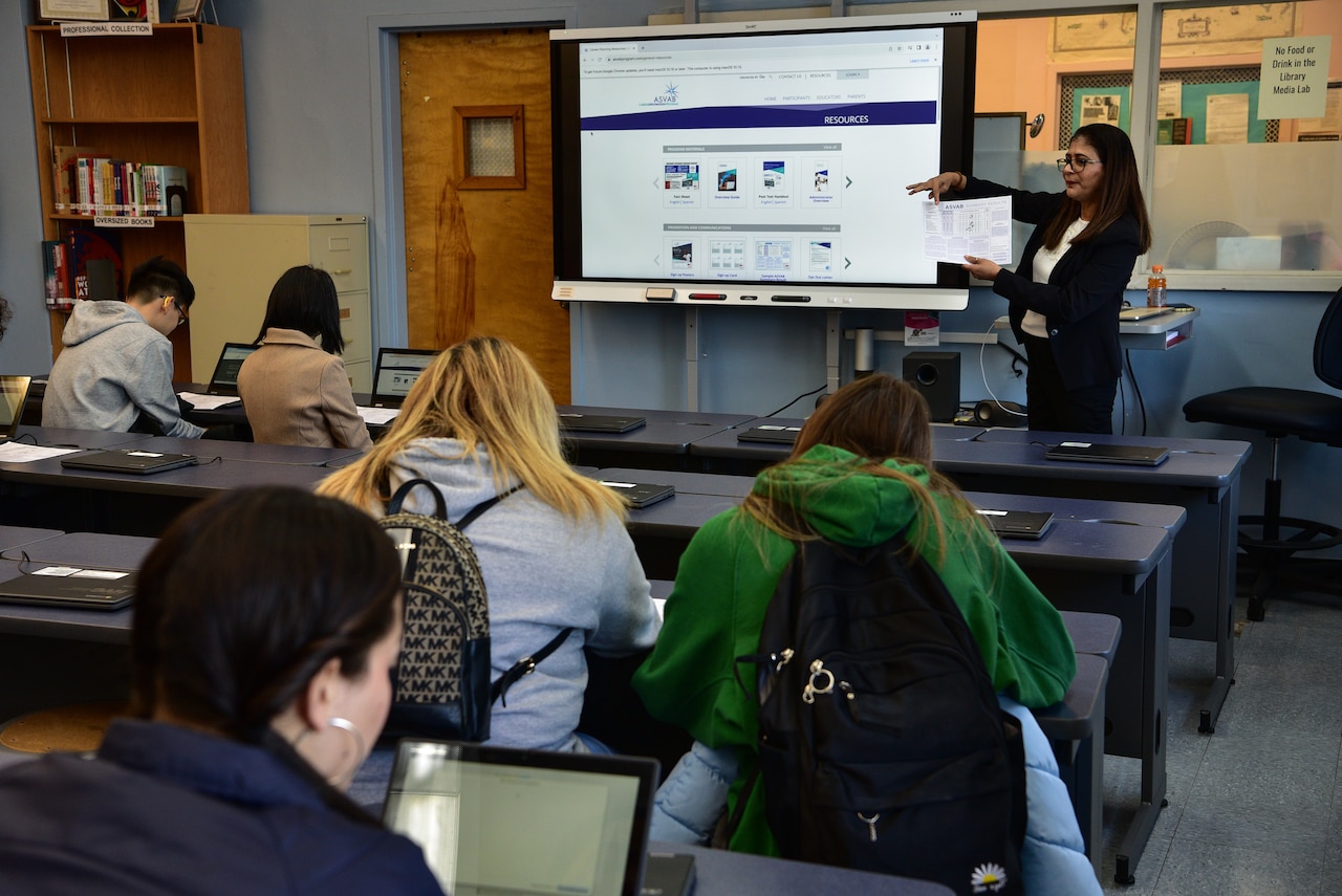 A person holds up a piece of paper while standing at the front of a classroom.