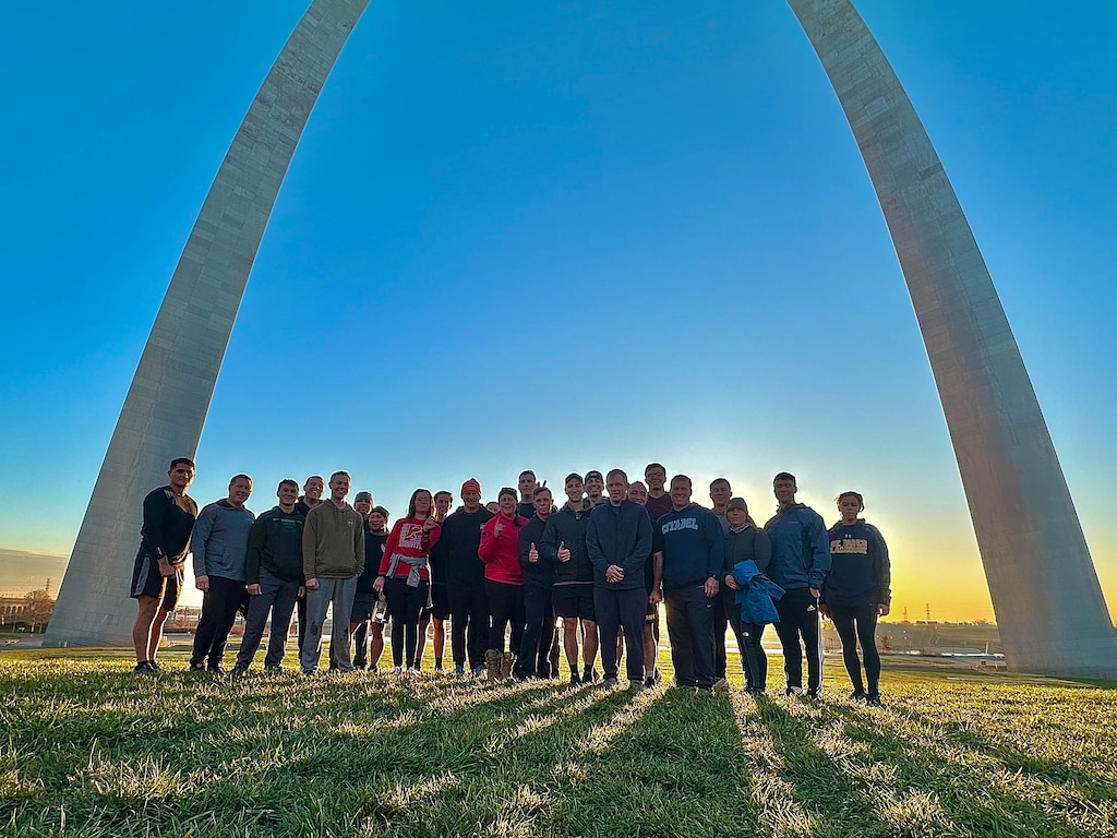 Several people in athletic clothing stand under the Arch with grass in the foreground and blue sky in the background.