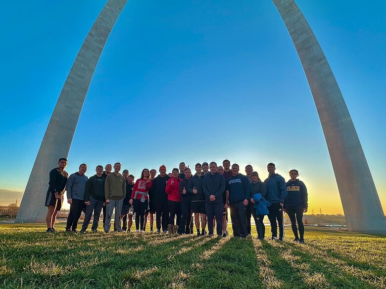 Several people in athletic clothing stand under the Arch with grass in the foreground and blue sky in the background.