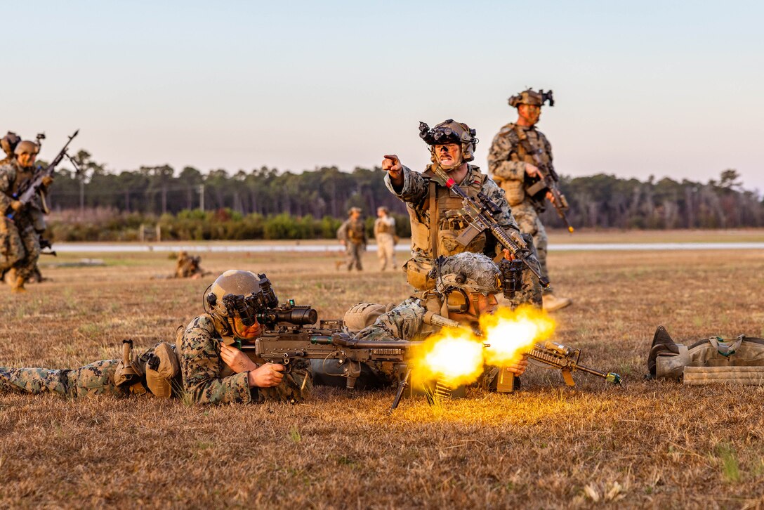 Two Marines lie on the ground while firing weapons as a third points while kneeling near them.