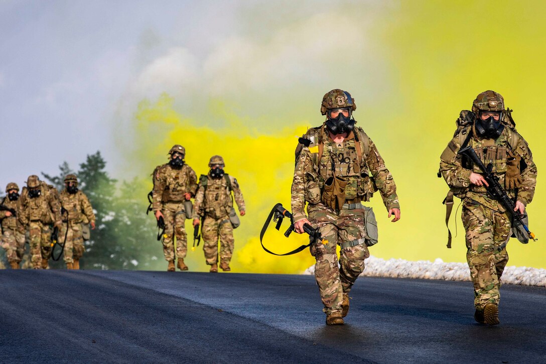 Soldiers walk down a street as yellow smoke rises in the background.