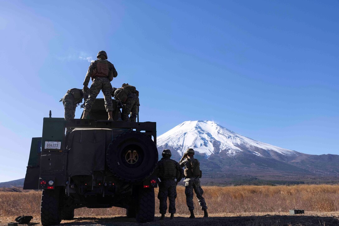 Marines stand on top of and next to a military vehicle facing a mountain peak.