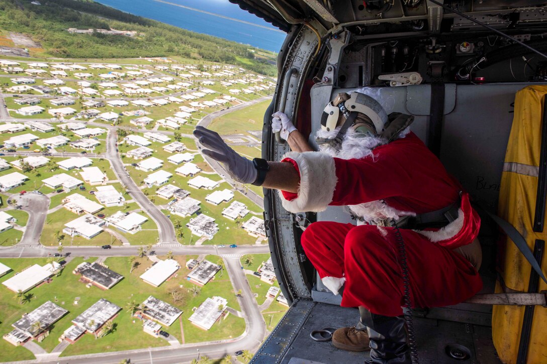 A sailor, dressed a Santa Claus, squats while waving out of the open door of an airborne helicopter to a community of homes.