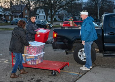 Outside in parking lot a pickup truck has Christmas packages unloaded by four people including DLA Disposition Services Director Mike Cannon. the sky is dark indicating it took place early in the morning. the packages from the truck are loaded onto a flat bed hand cart.