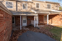 Photo of the front of two condominium homes with brick facades and tan sidings. There are brown fall leaves on the lawn and paved driveway.