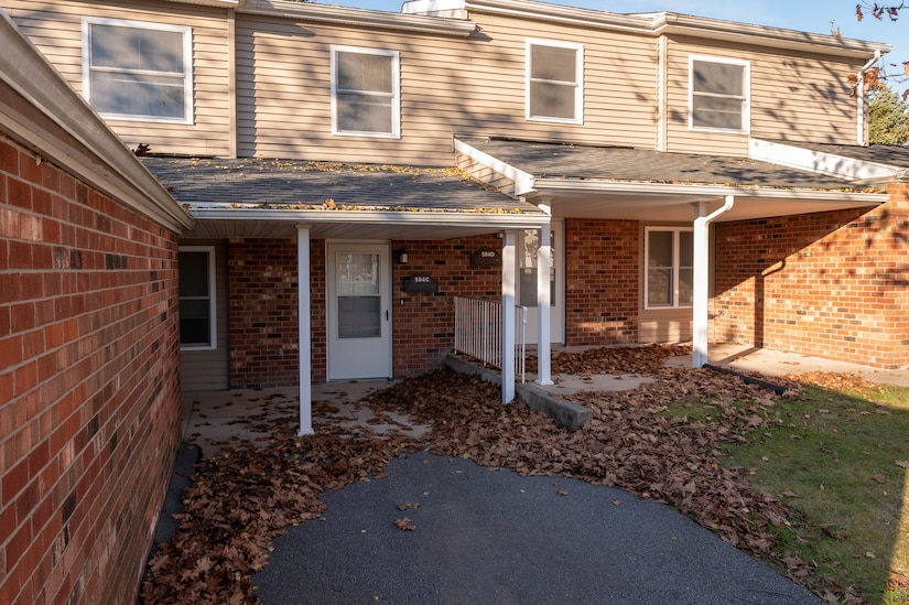 Photo of the front of two condominium homes with brick facades and tan sidings. There are brown fall leaves on the lawn and paved driveway.