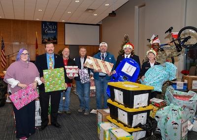 Eight people stand together looking at the camera holding Christmas gifts. One man is in an Army uniform and is holding a child's bike up in the air.