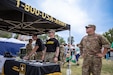 Four Soldiers standing underneath an Army tent host a recruitment booth in a field.