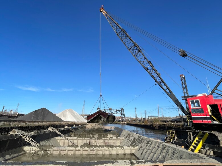 A red crane drops dredged sediment in a scow floating on a river channel.