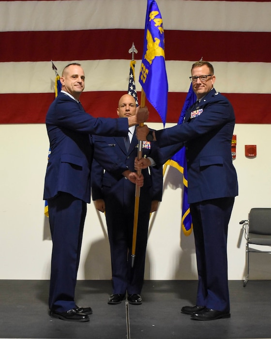 Two men of equal height in Air Force blue service dress uniforms grip the staff of a flag in front of a much shorter bald man.