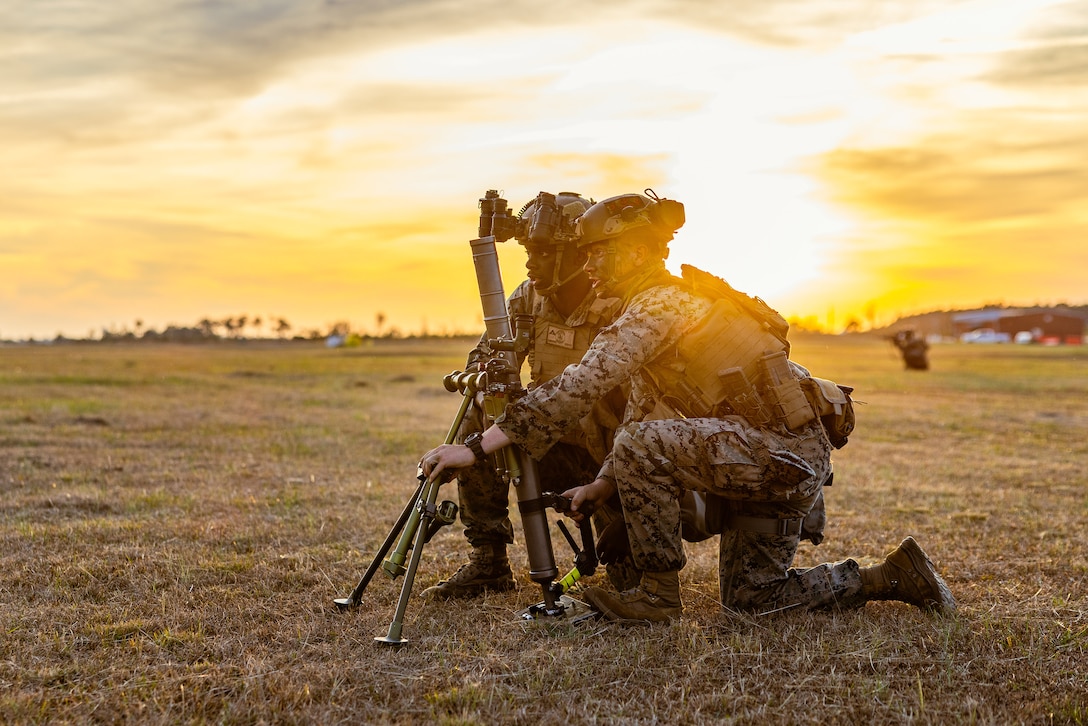 Marines kneel on a field to load a mortar with a low, bright sunrise in the distance.
