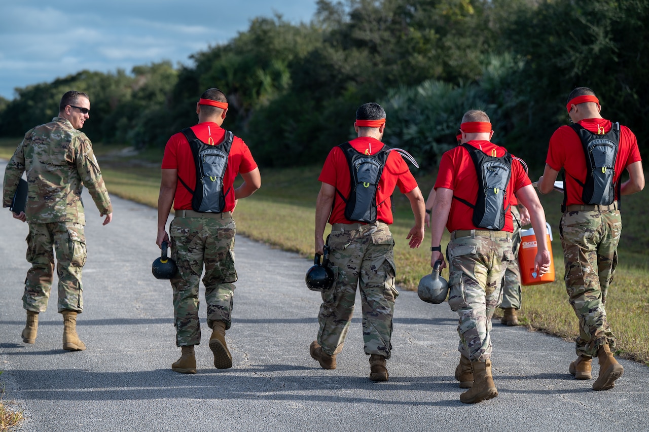 Guardians walk on pavement while carrying kettle bells.