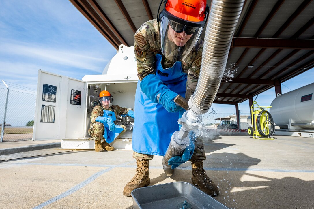 An airman wearing protective gear uses a tool to detach liquid nitrogen pipes as a sitting airman watches from the background.