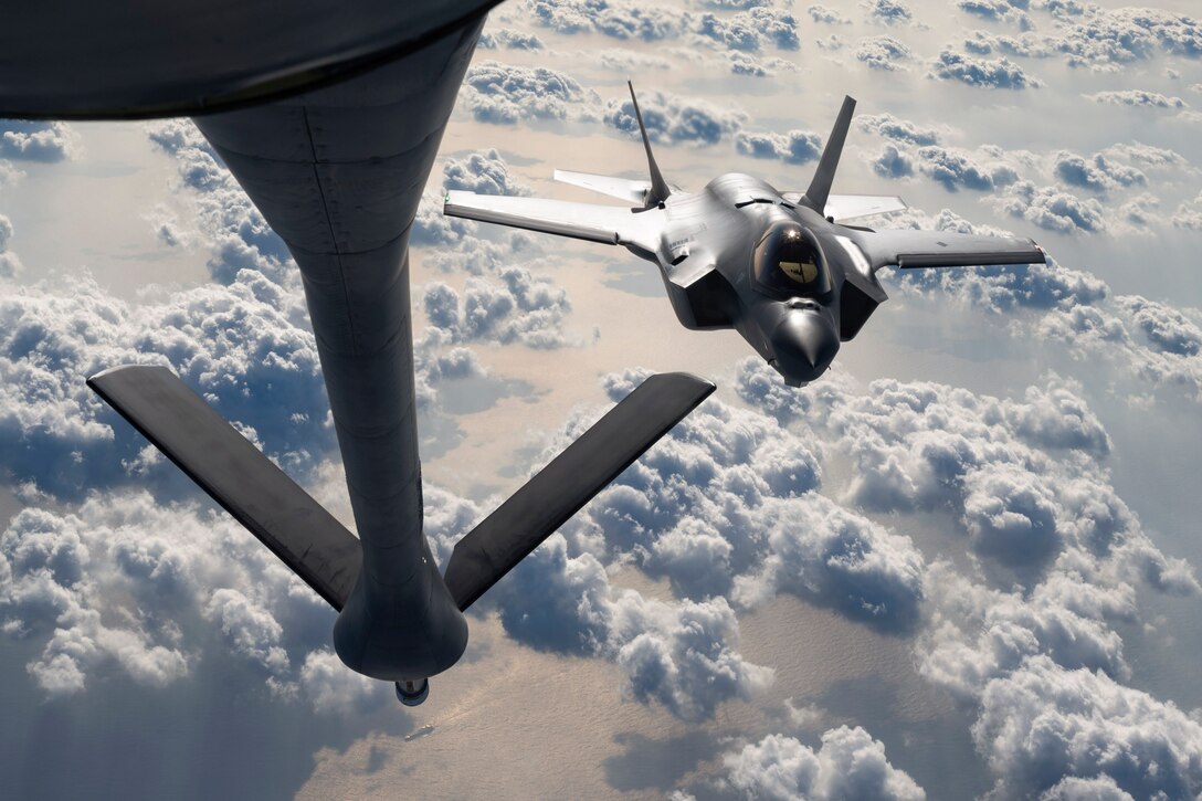 An aircraft’s boom sits to the left as another aircraft flies to the right above clouds and a body of water in the distance.