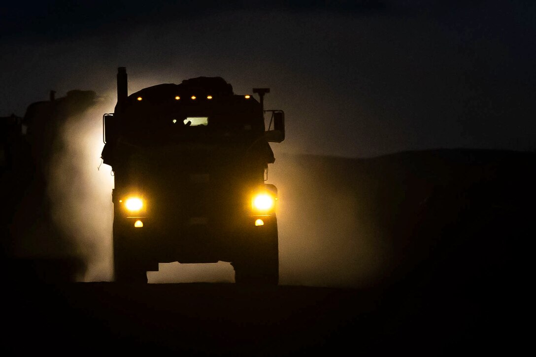 A military vehicle shown in silhouette drives down a road at night.