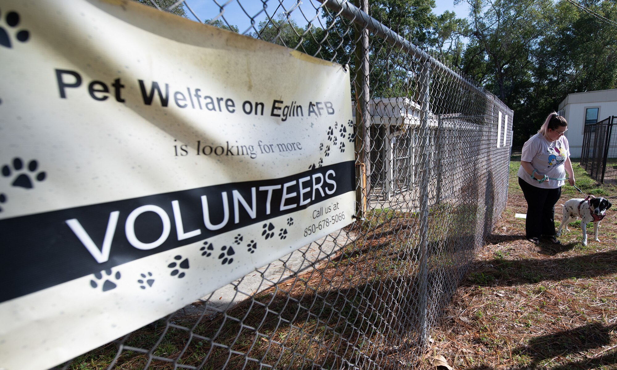 A sign reads, "Pet Welfare on Eglin AFB is looking for more volunteers." In the background is a volunteer walking a dog next to the fence with the sign.