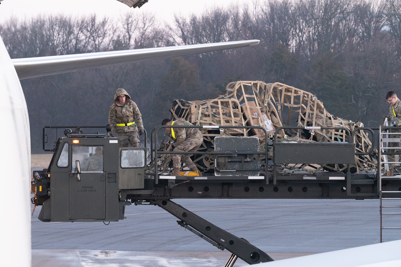 Service members stand next to a pallet of military equipment staged near a cargo plane.