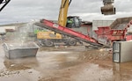 Glass flies out of an industrial shredder into a large garbage bin at a scrap yard.