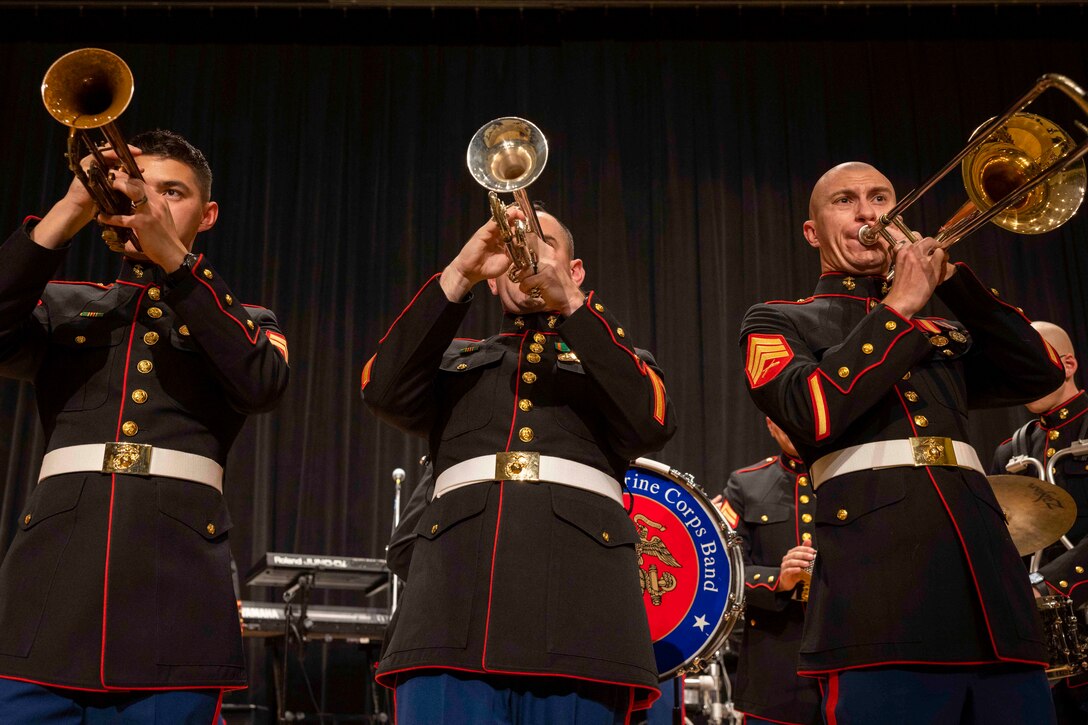 Two Marines play trumpets and another Marine plays a trombone while standing side by side on a stage.