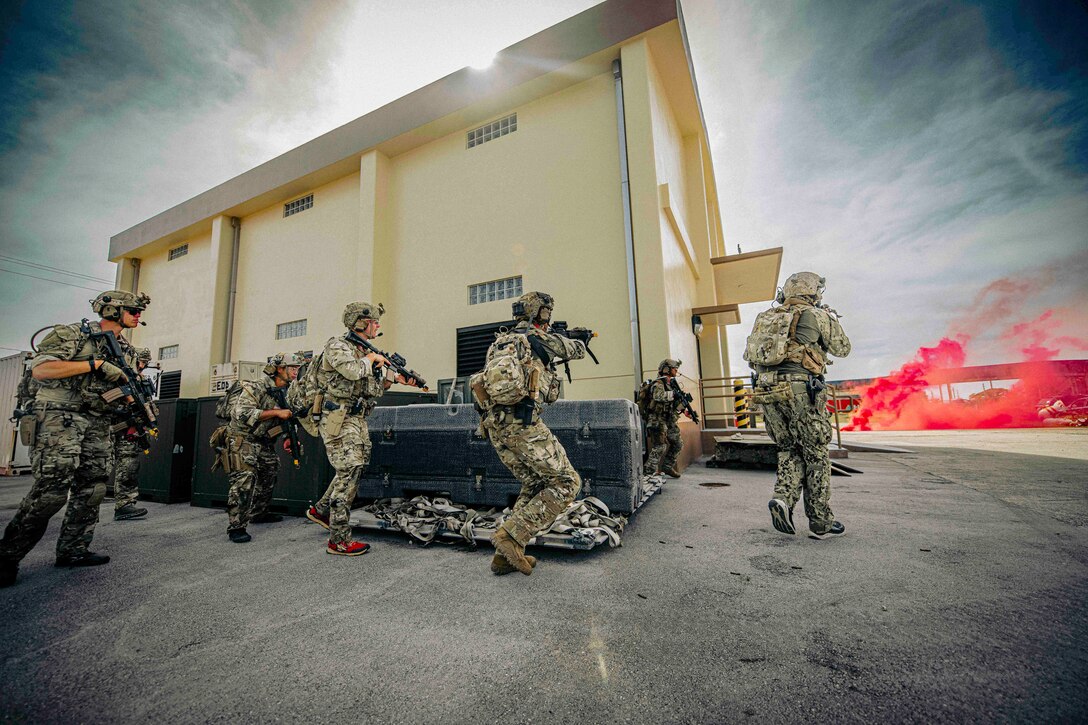 Sailors and Marines aim weapons while running next to a yellow building toward clouds of red smoke.