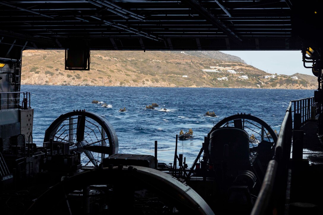 .S. and Greek marines ride in small boats in a body of water with mountains in the background as seen through the hangar bay of a ship.