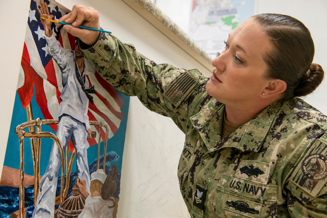 A sailor tilts their head while painting a mural of sailor aboard a ship with an American flag in the background.