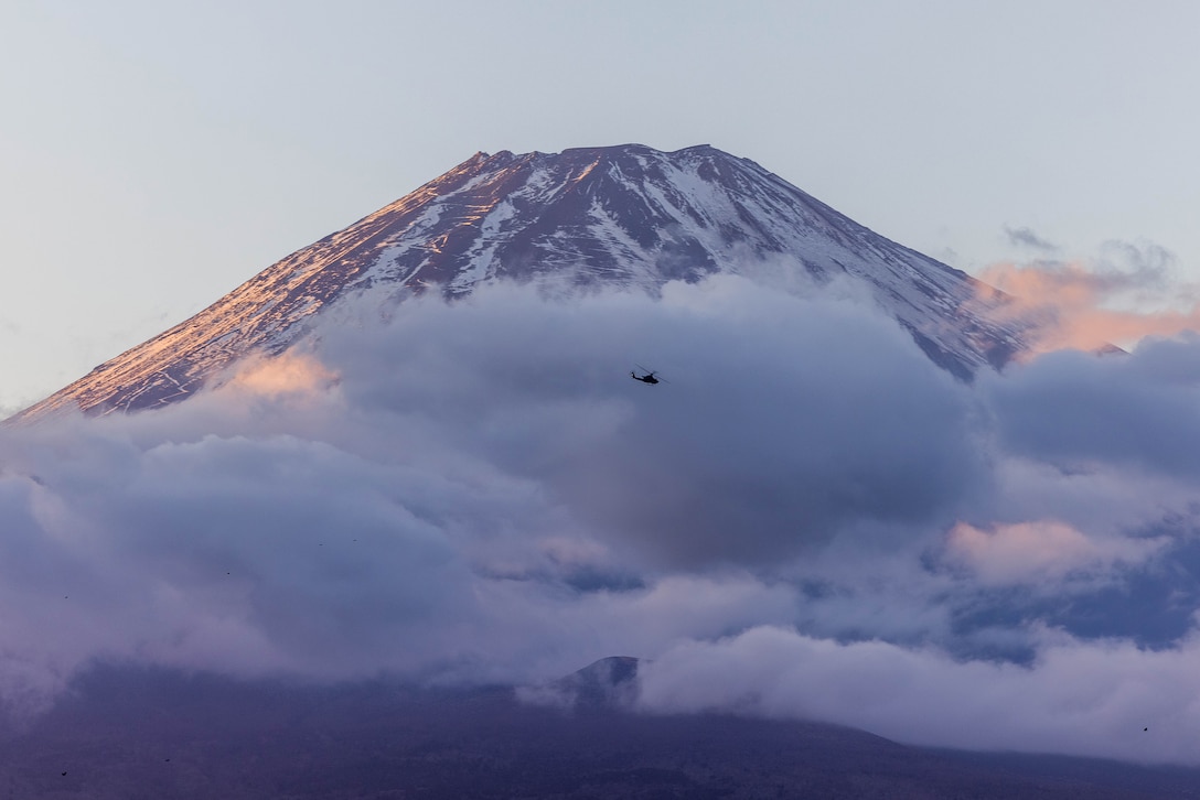 A military helicopter flies over clouds during daylight with a snowy mountaintop in the background.