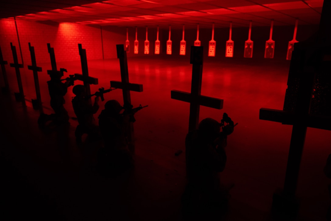 Airmen aim weapons at targets in an indoor shooting range. The room is illuminated by a red light.