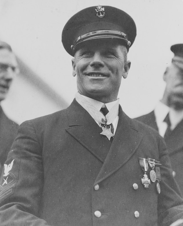 A man in a uniform and cap wears a medal around his neck and smiles.