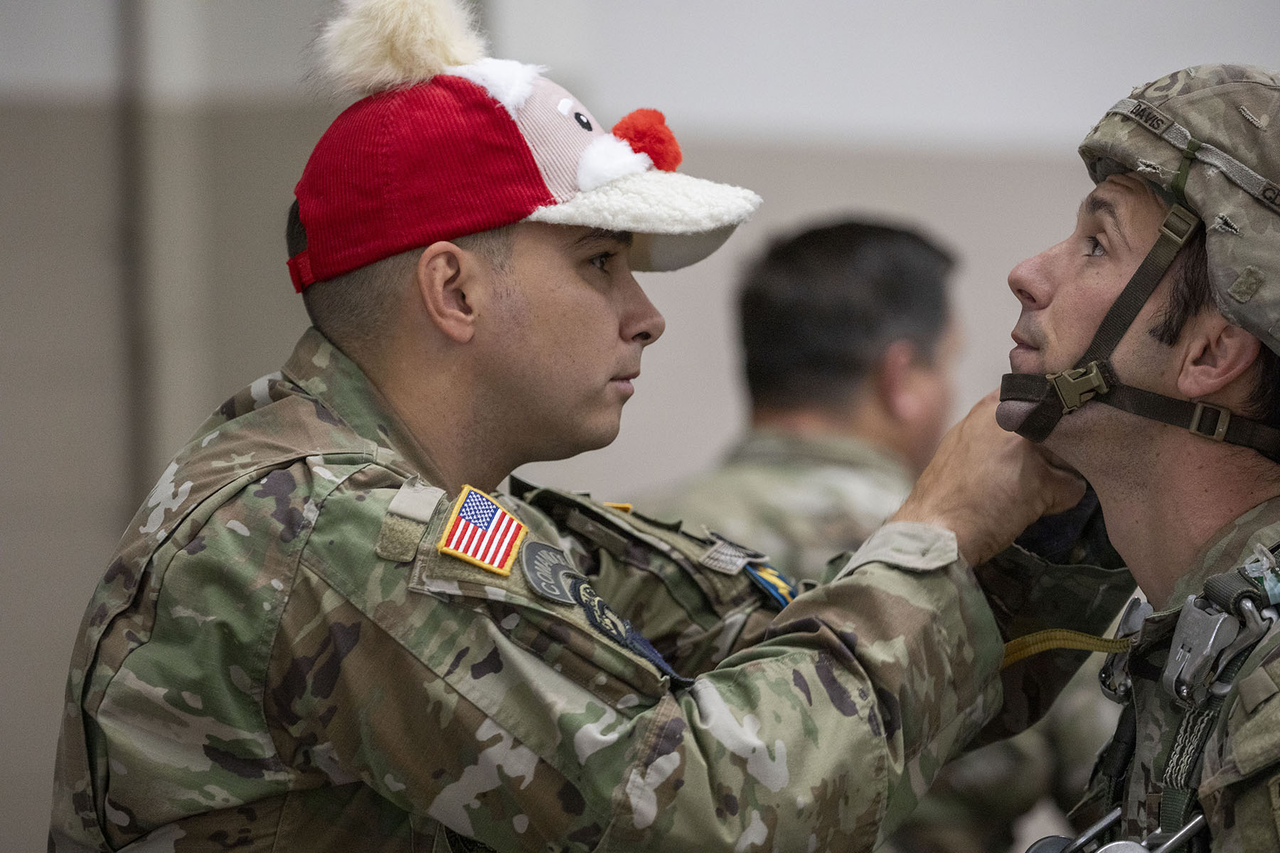 A soldier wearing a Santa baseball cap helps another soldier strap on a helmet. 