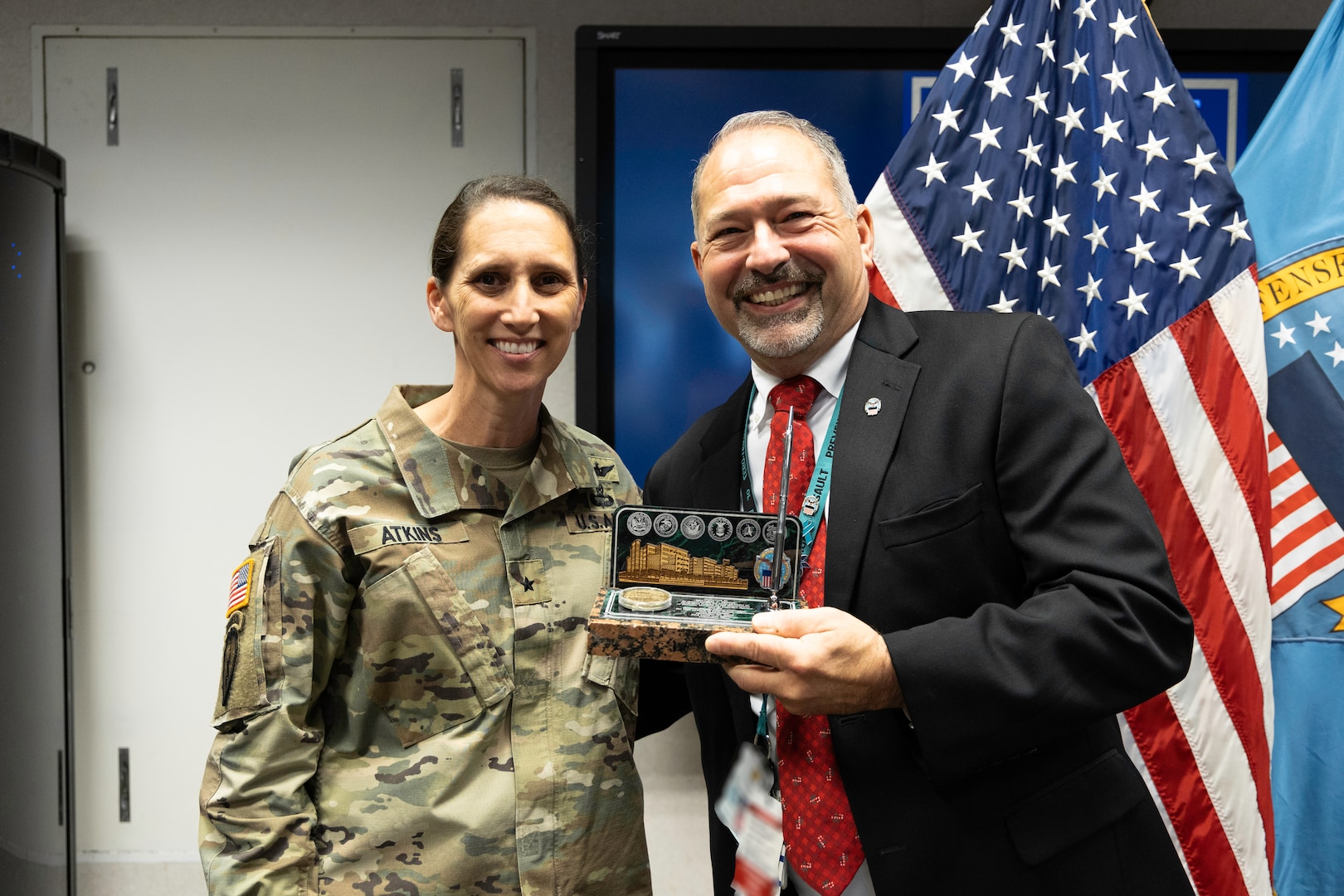 A light skinned woman with dark hair wearing a camouflage uniform smiles at the camera while giving a light skinned man, also smiling, with graying hair and beard wearing a dark suit and a red tie a stone desk set with engraving and a pen in a conference type room.