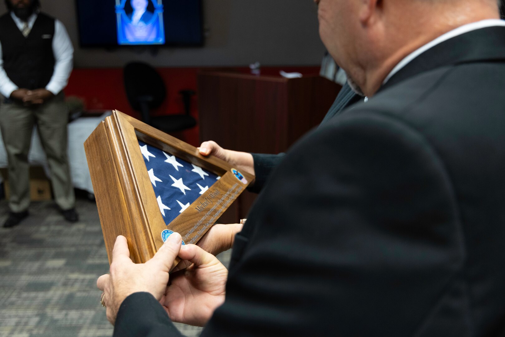 A light skinned man in a dark suit with graying hair looks at a U.S. Flag encased in a light brown box.