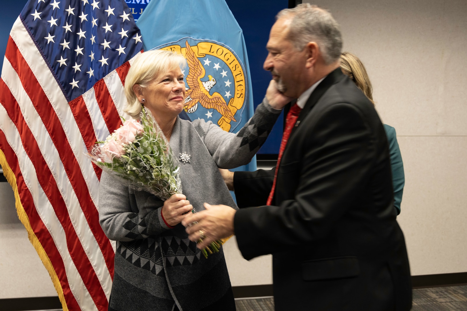 A light skinned man with graying hair in a dark suit with red tie hands pink flowers to a light skinned woman in a gray sweater in a conference type room.