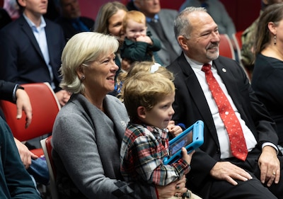 A light skinned man with graying hair and beard wearing a dark suit with a red tie laughs at a slideshow shown of his career in a conference type room. Next to him is a light skinned woman holding a small light skinned boy holding a iPad and a light skinned woman sits behind them holding a red-haired baby.