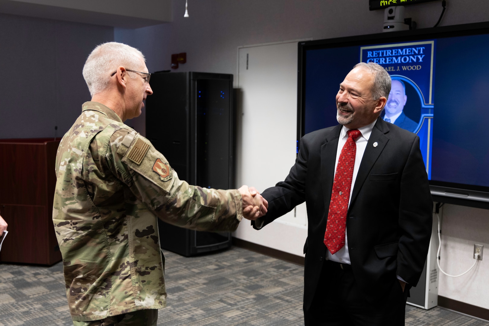 A light skinned man in a camouflage uniform with graying hair and glasses shakes the hand with another light skinned man with graying hair and beard wearing a dark suit with a red tie in a conference type room.