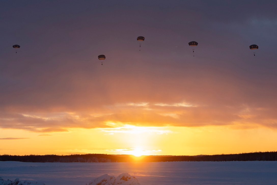 Service members descend in parachutes toward snowy ground with clouds and a low sun behind them.