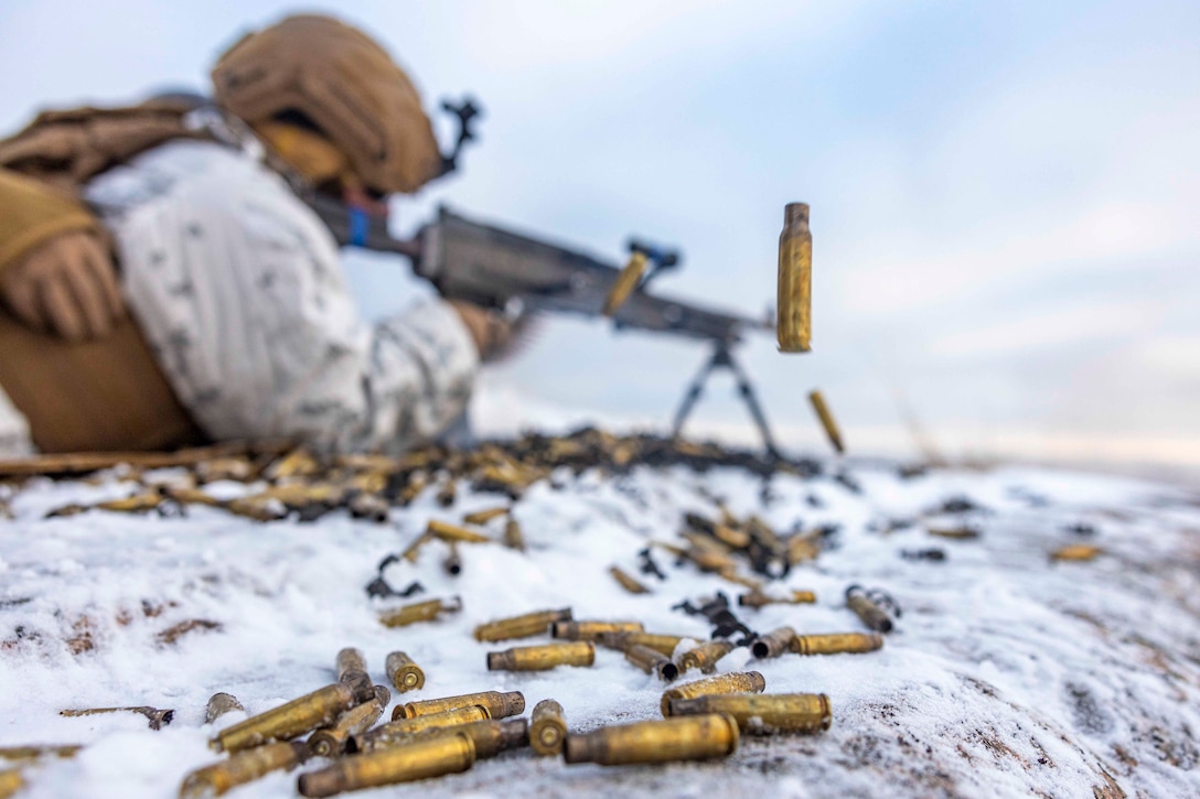 A Marine fires a weapon while lying on a snow-covered ground.