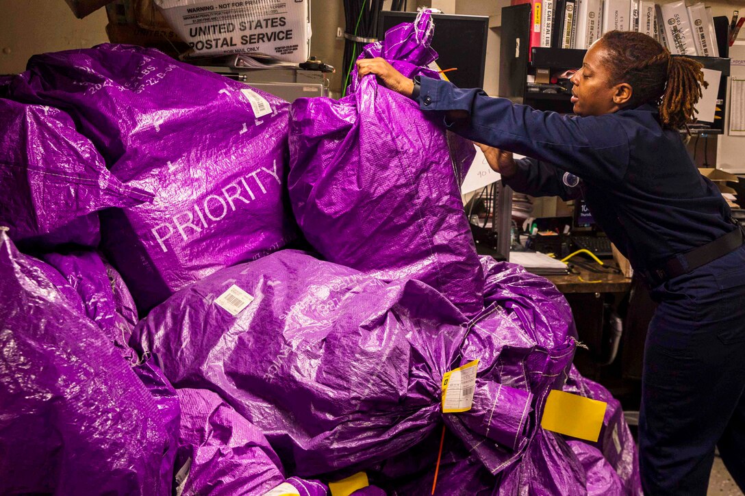 A sailor stacks purple bags marked "priority" in an office-like area.