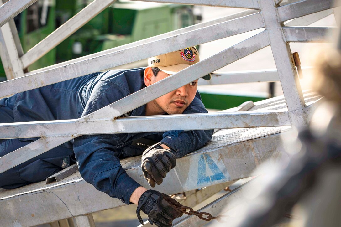 A sailor laying on a ramp holds a chain as seen through a metal gate.