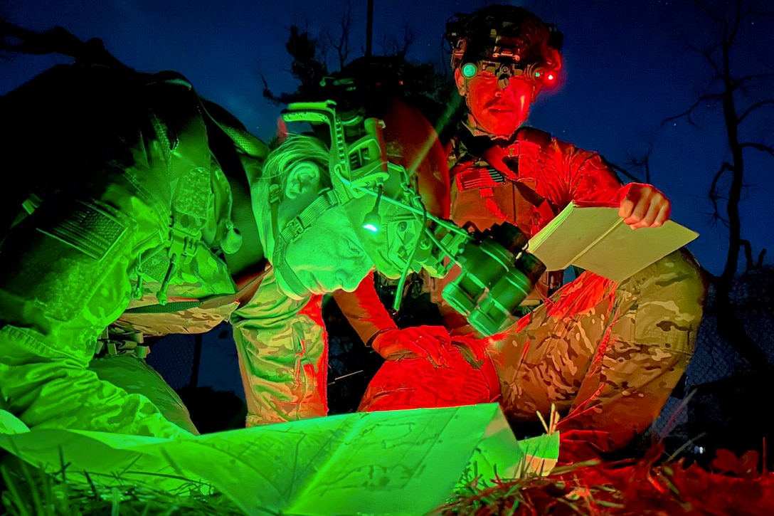An airman wearing night vision goggles looks at a map as a fellow kneeling airman holds a book open in the dark illuminated by red and green lights.