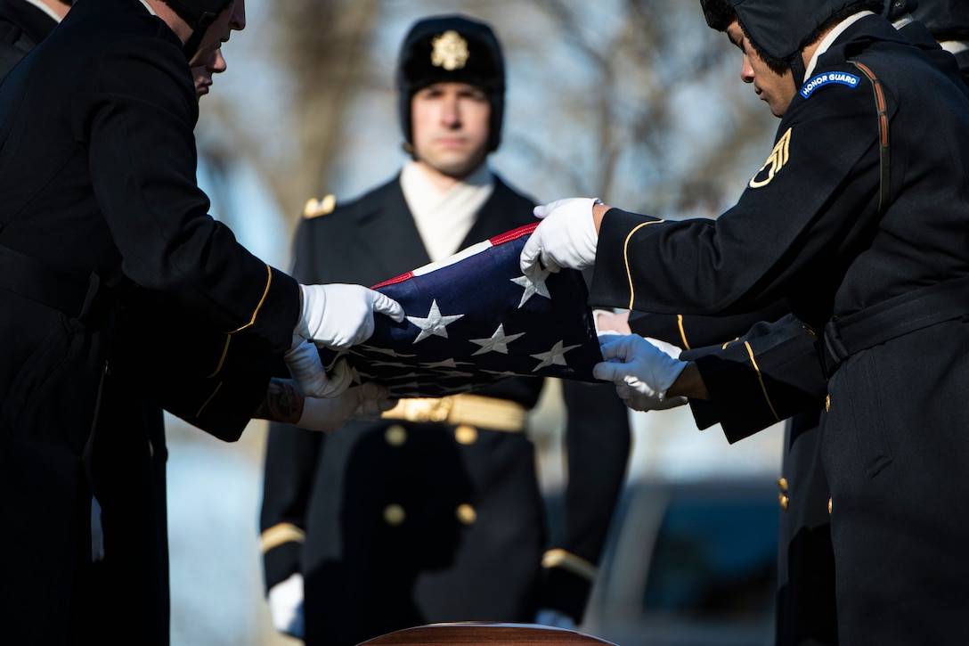 The American flag is folded as a soldier stands at attention during a funeral service.