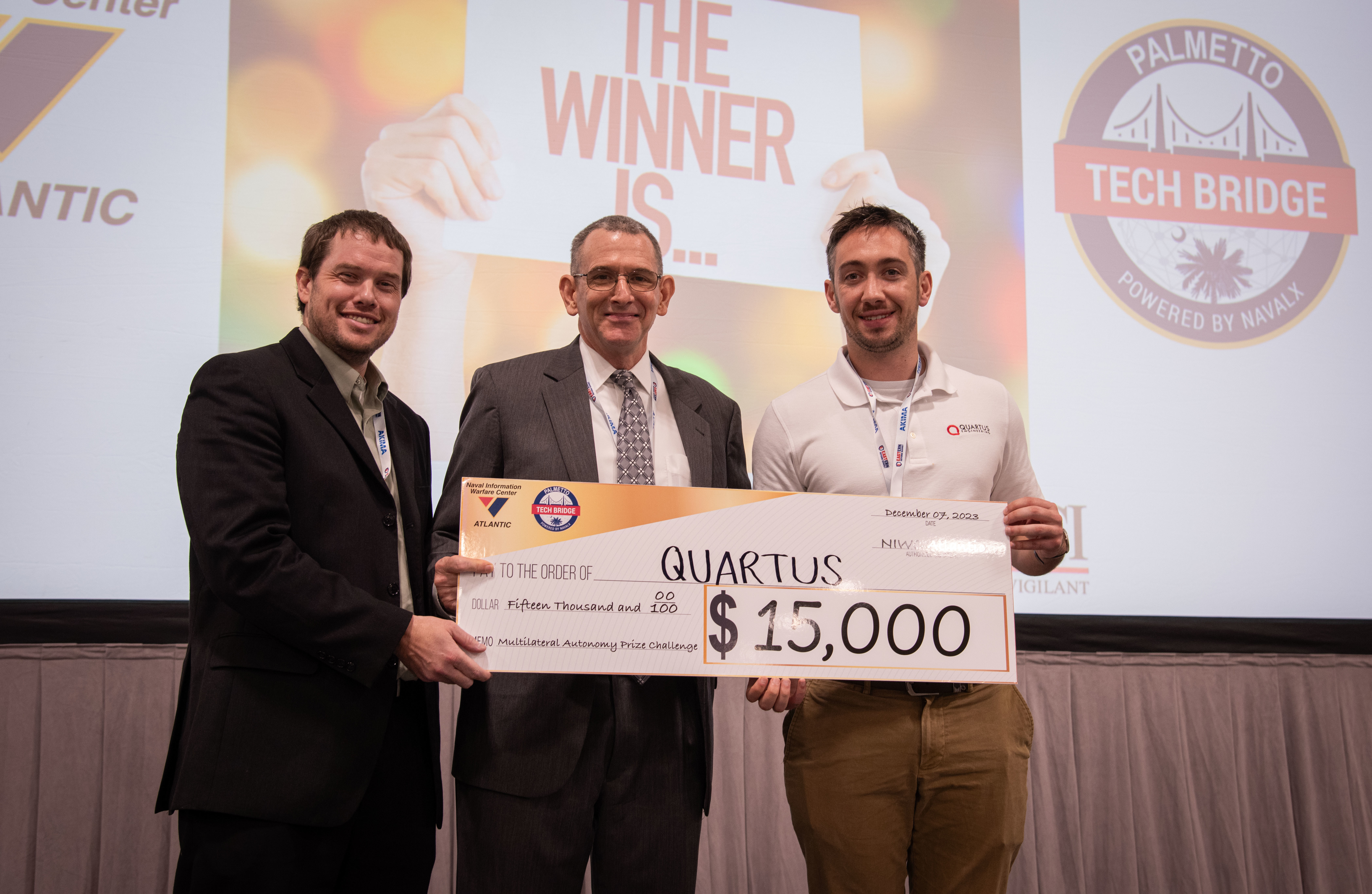 Mr. Peter C. Reddy, middle, executive director of Naval Information Warfare Center (NIWC) Atlantic, and Dr. Luke Overbey, left, NIWC Atlantic’s senior scientific technical manager for autonomy, present a $15,000 check to Quartus on Dec. 7 for taking second place in the Multilateral Autonomy Prize Challenge at the 2023 Eastern Defense Summit.