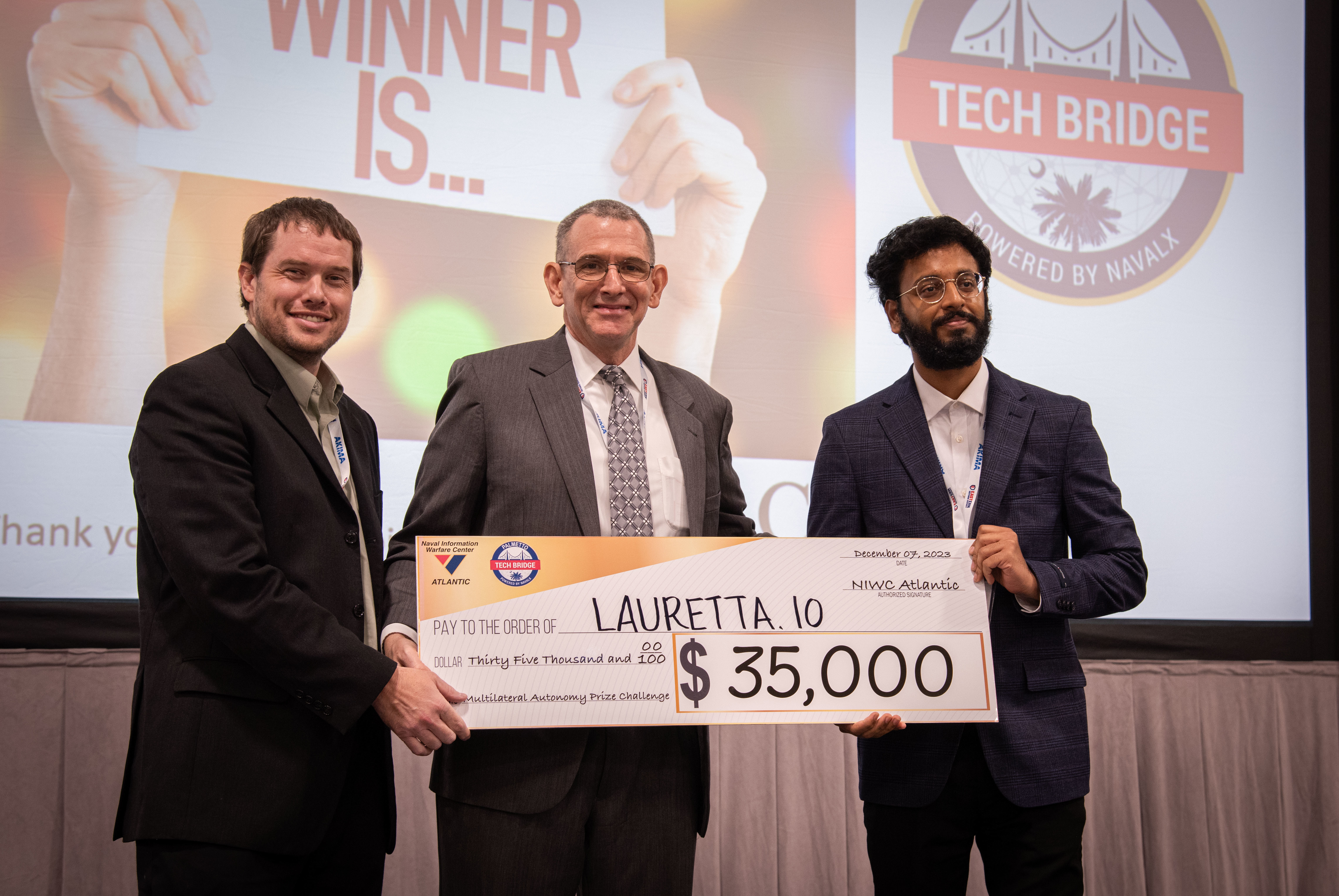 Mr. Peter C. Reddy, middle, executive director of Naval Information Warfare Center (NIWC) Atlantic, and Dr. Luke Overbey, left, NIWC Atlantic’s senior scientific technical manager for autonomy, present a $35,000 check to Lauretta.io on Dec. 7 for winning first place in the Multilateral Autonomy Prize Challenge at the 2023 Eastern Defense Summit.