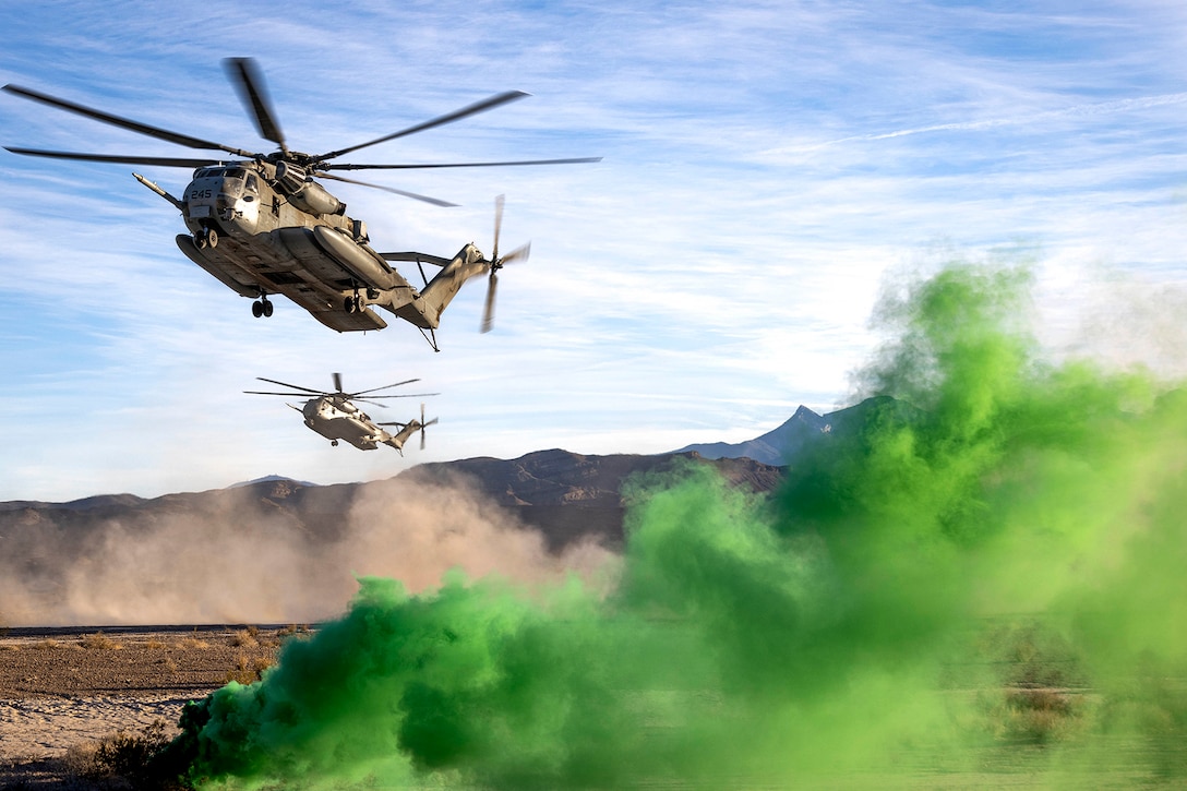 Marine Corps helicopters fly near the ground as green smoke is shown in the lower right of the photo.