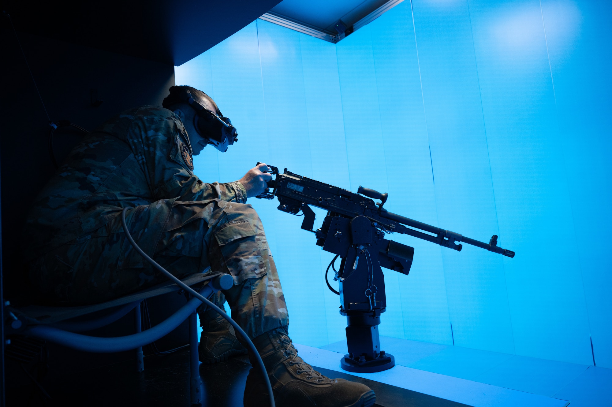 A man in military uniform sits in a blue simulator room as he sees a training scenario through virtual reality goggles and holds a training weapon towards the simulation.