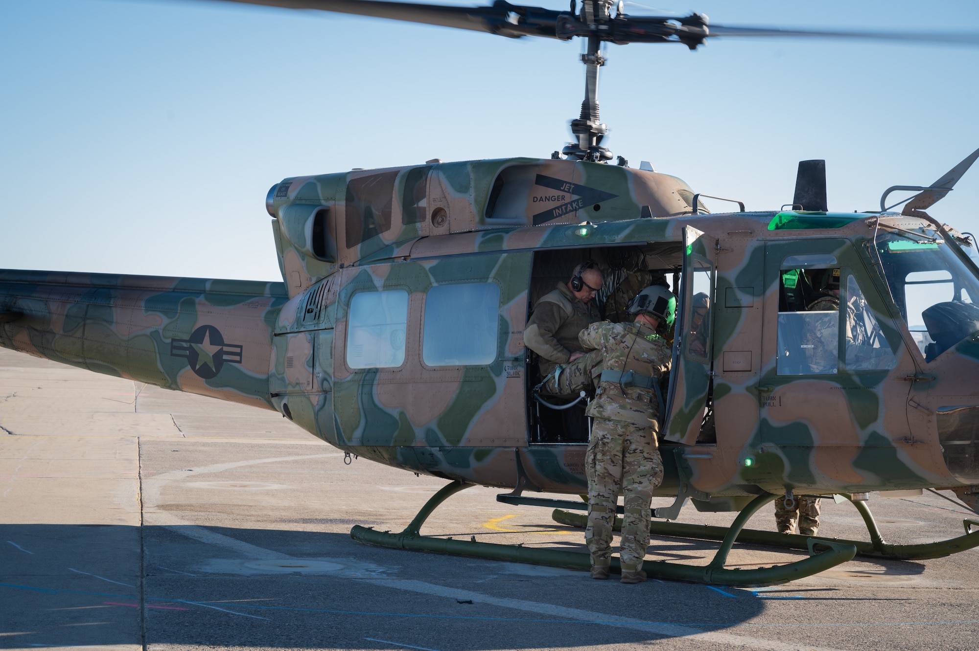 Two people in military uniform prepare for flight takeoff on a camouflage-painted helicopter.