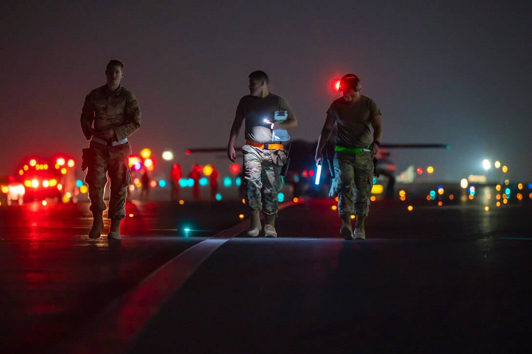 Three airmen walk next to one another in the dark in front an aircraft illuminated by colorful lights.