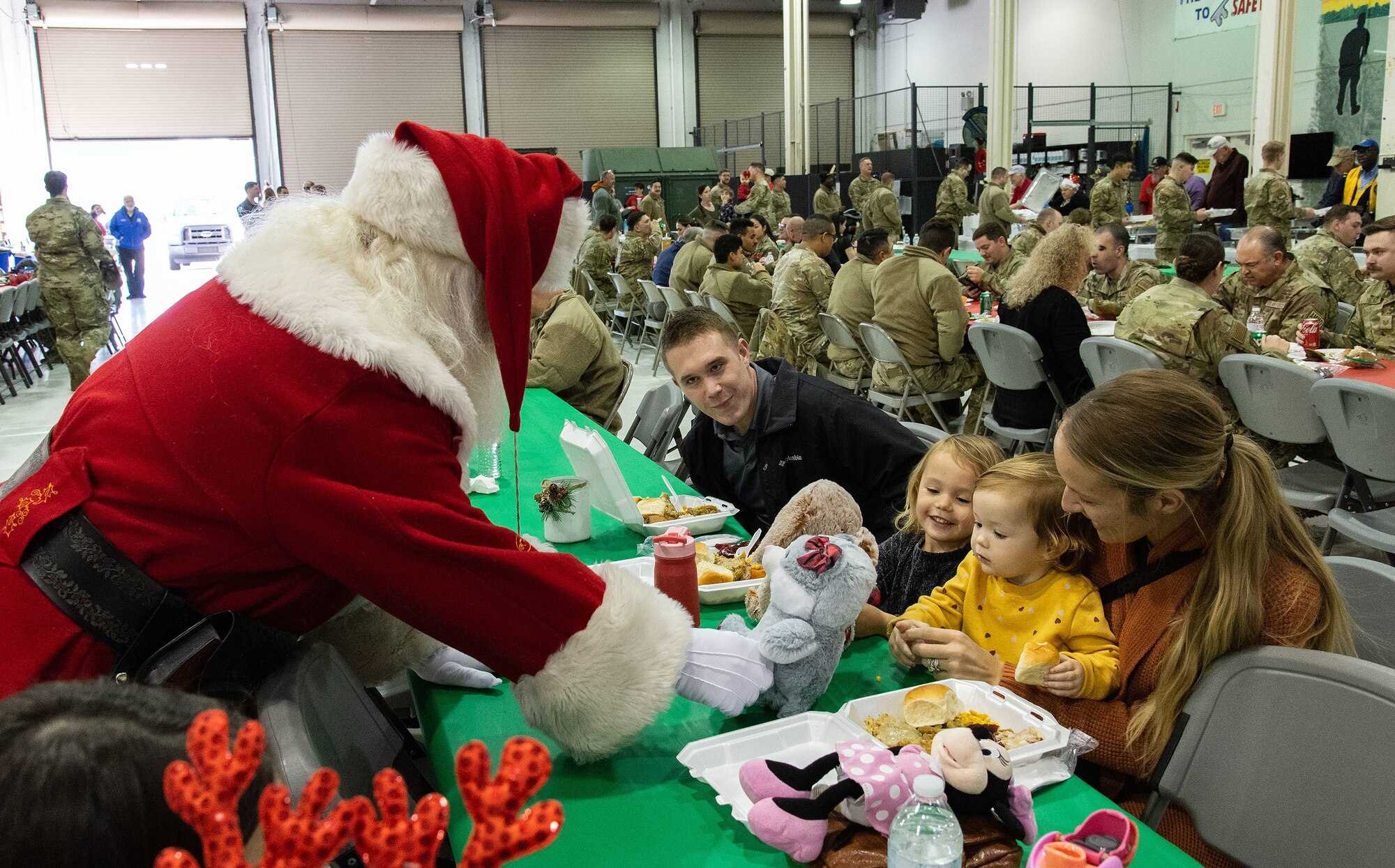 Greg ‘Santa’ Miller hands a stuffed animal to Myla MacIntyre, as her older sister Ava, watches while attending the Operation Feed the Troops meal at Dover Air Force Base, Delaware, Dec. 13, 2023. The two are the daughters of U.S. Army Sgt. 1st Class Lawrence MacIntyre, center, University of Delaware Army ROTC, Newark, Delaware, and his wife Jalena, right, who brought their children to the day meal and to meet Santa. (U.S. Air Force photo by Roland Balik)