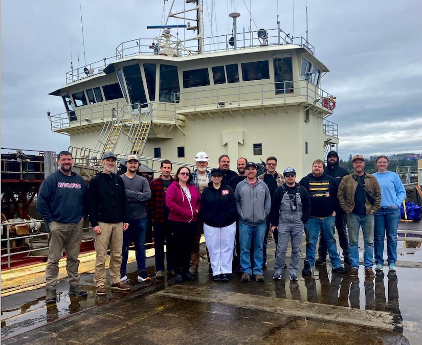 A crew of 16 people stand in front of the dredge vessel, the Yaquina.