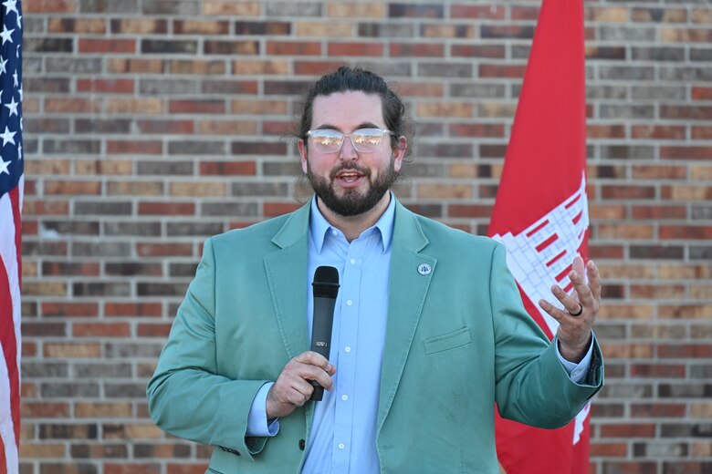 Man stands at podium in teal blazer with glasses and long hair.